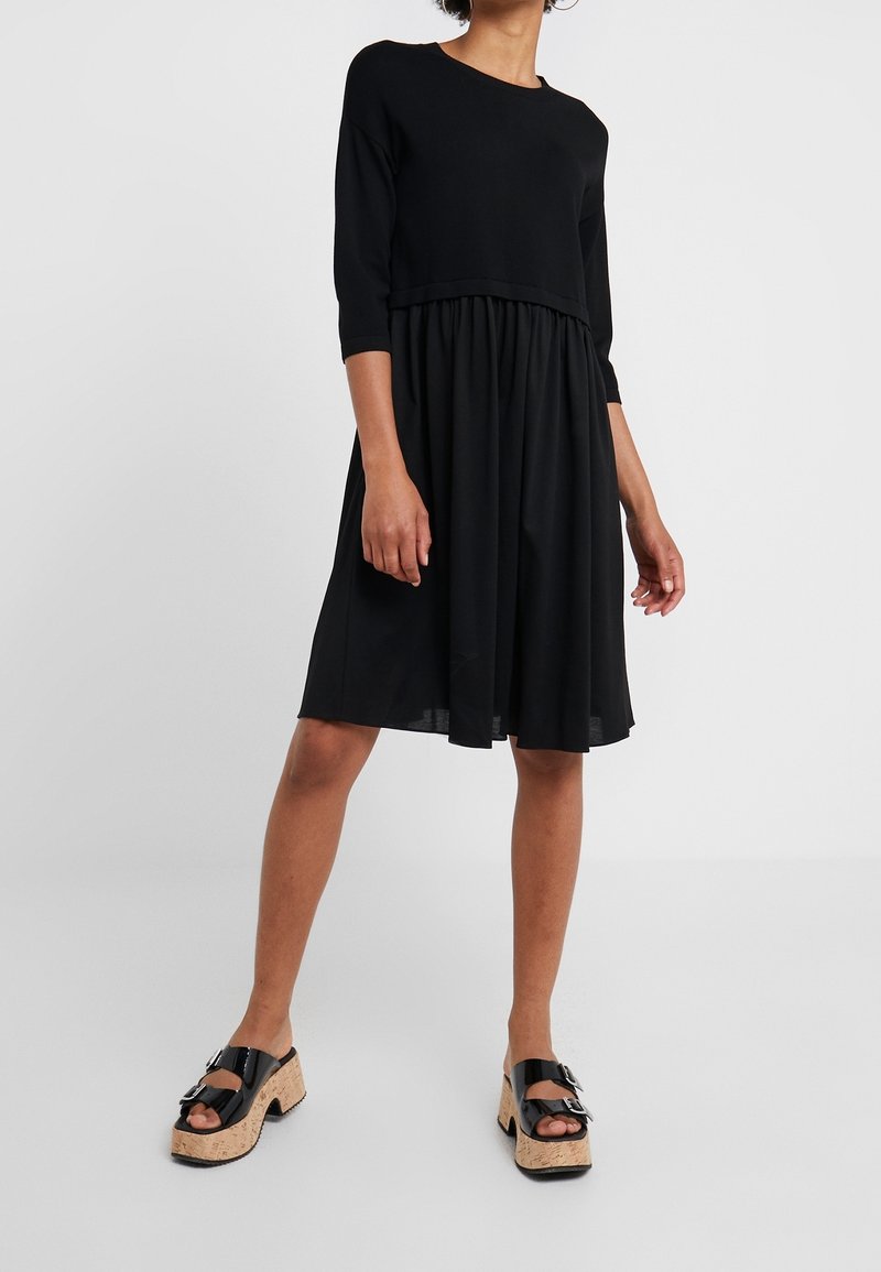 Femme portant une robe noire à manches trois-quarts avec une jupe froncée et des sandales compensées noires avec semelles en liège.