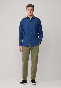 Camisa de botones de manga larga azul, pantalones cargo verdes y zapatillas negras, de pie contra un fondo sencillo, con las manos en los bolsillos.