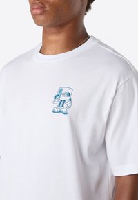 Mand iført en hvid T-shirt med en lille blå tegneseriefigur på brystet, der holder en kube med teksten "The North Face."