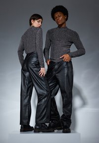 Dos modelos están sobre una plataforma, vestidos con tops de manga larga brillantes, pantalones negros de cuero sintético y botas negras, con un fondo gris.