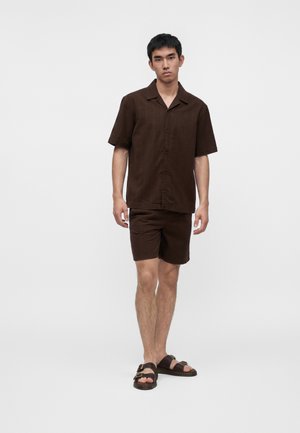Junger Mann steht, trägt ein dunkelbraunes kurzärmeliges Hemd mit Knöpfen, passende Shorts und braune Sandalen vor einem einfarbigen weißen Hintergrund.