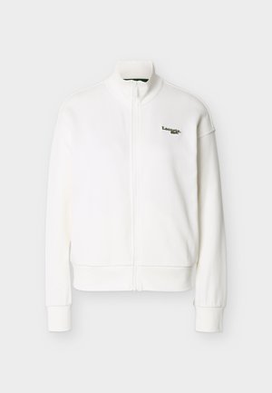 Jachetă albă cu fermoar, guler înalt, mâneci lungi, manșete riburate și un mic logo Lacoste verde pe partea stângă a pieptului.