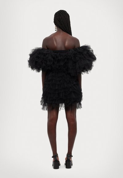 Zwarte off-shoulder jurk van gelaagd tule, met gerimpelde mouwen en een aangesloten silhouette, aangevuld met zwarte platformhakken.