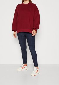 Sweat-shirt oversize bordeaux avec un col rond et un logo brodé, associé à un jean foncé et des baskets blanches avec des accents rouges.
