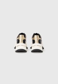 Een paar witte sneakers met zwarte en gouden accenten, gezien vanuit de achterkant tegen een effen witte achtergrond.