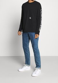 Černé tričko s dlouhým rukávem a malým bílým logem na hrudi, s nápisem "CALVIN KLEIN JEANS" vytištěným bílou barvou na pravém rukávu. V kombinaci s modrými džíny a bílými teniskami.