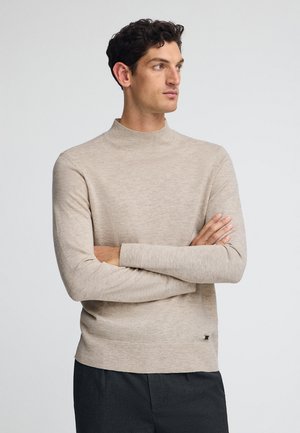 Man with short curly hair wearing beige long-sleeve sweater and dark pants, standing with arms crossed, looking to the side.