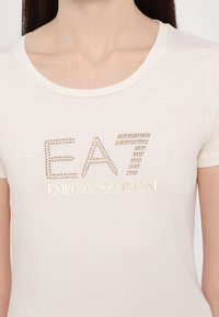 Kriešanas īsajiem piedurknēm T-krekls ar "EA7 EMPORIO ARMANI" zeltītās spalvotās burtiem, ar gludu tekstūru un noapaļotu kakla izgriezumu.