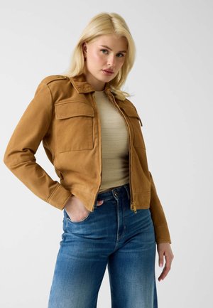 Blonde vrouw die een beige suède jas draagt over een beige top en een hoge taille blauwe jeans, poserend met één hand in haar zak tegen een witte achtergrond.
