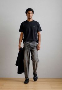 Μαύρο κοντομάνικο μπλουζάκι με μικρό λογότυπο στο στήθος, συνδυασμένο με ανοιχτό γκρι φαρδύ τζιν και μαύρα παπούτσια, κρατώντας ένα μαύρο μπουφάν.