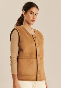 Braune Weste mit Wildleder-Oberfläche, runder Hals Ausschnitt, Knopfverschluss vorne, zwei aufgesetzte Taschen und ärmelloses Design. Schmale Passform.