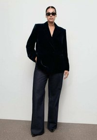 Mujer con gafas de sol oscuras, vistiendo un blazer de terciopelo negro, pantalones de pierna ancha oscuros y zapatos negros de punta, de pie contra una pared lisa.