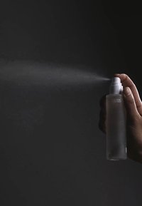 Przezroczysta butelka ze spryskiwaczem w białym kolorze, trzymana w ręku, uwalniająca delikatną mgiełkę na ciemnym tle. Matująca, szklana tekstura.