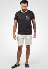 Camiseta negra de manga corta con un bolsillo con un patrón de hojas verdes, combinada con pantalones cortos gris claro y zapatos oscuros de tipo slip-on.
