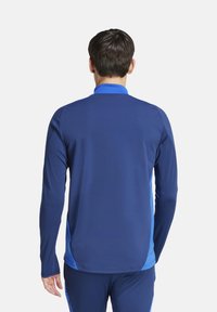 Maglietta sportiva a maniche lunghe blu con colletto e pannelli laterali a contrasto. Realizzata in un materiale liscio e elastico per comfort e libertà di movimento.