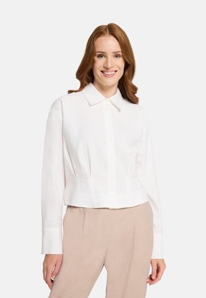 Smiling woman with long red hair wearing a white buttoned shirt and beige pants against a plain background.