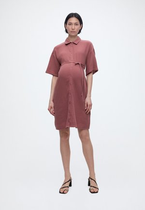 Έγκυος γυναίκα όρθια, φορώντας ένα κουμπωτό φόρεμα με μήκος έως το γόνατο σε σκουρό ροζ απόχρωση, με κοντά μανίκια και μαύρα σανδάλια με τακούνι.