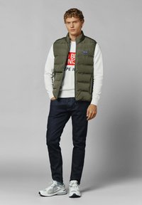 Khaki puffer vest over een wit sweatshirt met rode en blauwe tekst, gecombineerd met donkere jeans en grijze sneakers. Glad materiaal, aansluitend ontwerp.