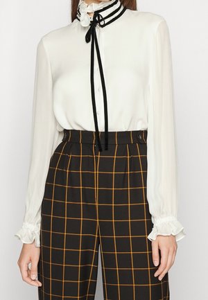 Blusa blanca de manga larga con cuello y puños con volantes, lazo negro, combinada con pantalones negros de cintura alta con patrón de cuadros naranjas.