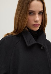 Schwarzer Wollmantel mit hohem Kragen, mit einem Knopfverschluss und strukturiertem Stoff. Das Design ist minimalistisch und klar strukturiert.