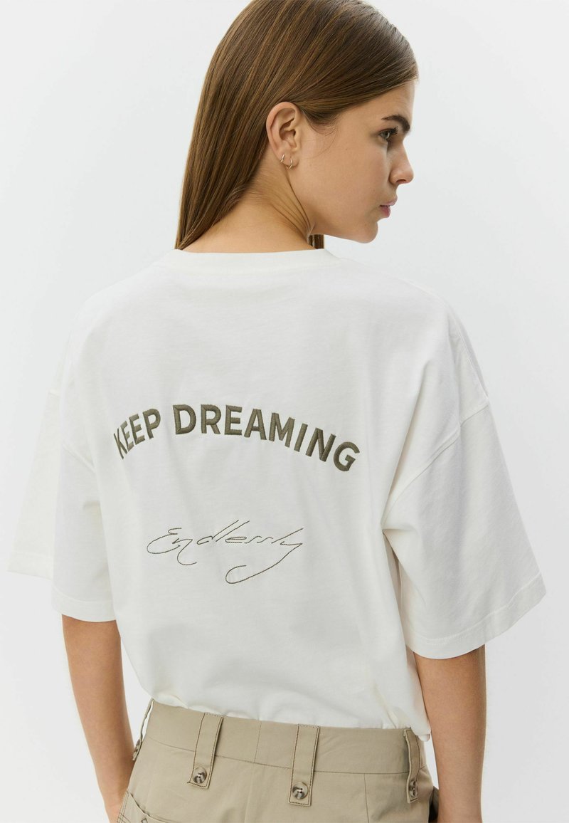 Tricou alb din bumbac cu mâneci scurte, având brodat cu verde măsliniu mesajul „KEEP DREAMING” și cu scris cursiv cuvântul „Endlessly” dedesubt.