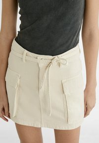 Bela denim mini krilo s pasom za zavezovanje, ki ima dva sprednja žepa in gladko teksturo. Zgornji del je temno siv ribičasti top.