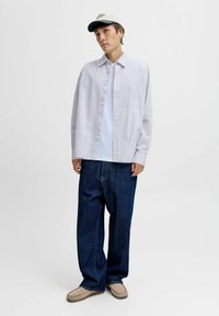 Ung mand iført hvid stribet skjorte over hvid t-shirt, mørkeblå brede bukser, beige slip-on sko og beige baseball cap mod hvid baggrund.