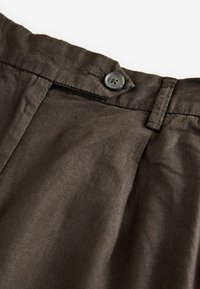 Ceinture de pantalon marron foncé avec un bouton noir et une boucle de ceinture visible sur un tissu texturé.
