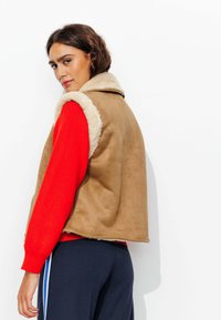 Gilet en faux daim marron avec col en polaire beige, associé à une chemise à manches longues rouge vif et un pantalon sombre avec des bandes latérales.
