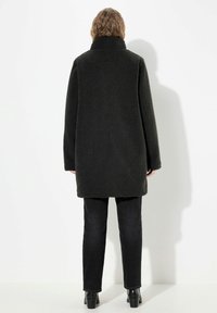 Cappotto in pile nero accogliente con colletto alto, vestibilità ampia e maniche lunghe. Abbinato a jeans scuri slim e stivaletti alla caviglia.