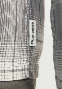 Grå rutigt skjorta med varierande linjebredd, etikett med "PRJCT-CORE" och en texturerad tyg. Synlig bomullsblandning.
