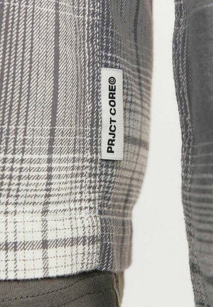 Chemise à carreaux grise avec des épaisseurs de ligne variées, étiquette portant "PRJCT-CORE", et un tissu texturé. Matériau en mélange de coton visible.