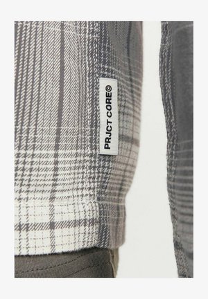 Camicia a quadri grigia con variazioni nello spessore delle linee, etichetta con "PRJCT-CORE" e un tessuto texturizzato. Materiale in misto cotone visibile.