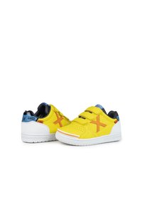 Zapatillas amarillas con suelas blancas y dos correas de velcro. Presentan material perforado y un diseño de "X" naranja en los lados. Forro negro.
