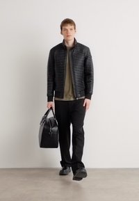 Melnā quilted jaka ar rāvējslēdzēju frontē, saskaņota ar zaļu augšu un melnām biksēm. Modelis tur melnu sporta somu, uzvilcis melnas kurpes.