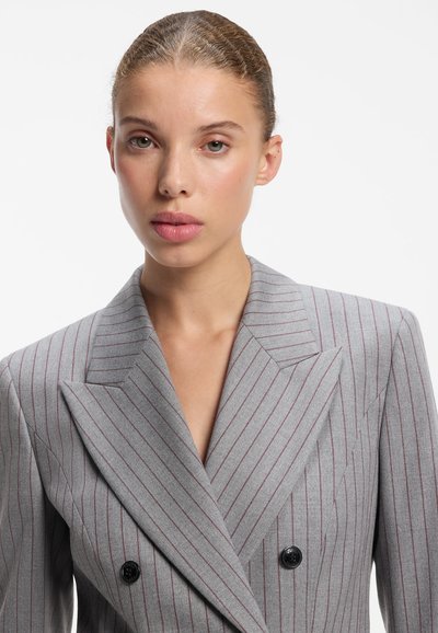 Jeune femme aux cheveux plaqués en arrière portant un blazer gris à fines rayures avec des boutons noirs sur fond blanc uni.
