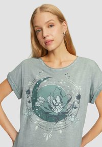 T-shirt en coton vert clair avec un graphique floral circulaire en teal et blanc, manches courtes et décolleté arrondi.