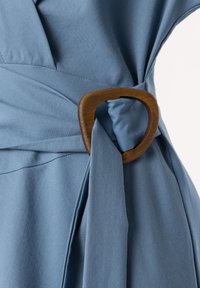 Article en tissu bleu présentant une finition texturée, cintré à la taille avec une boucle en bois créant un effet noué.