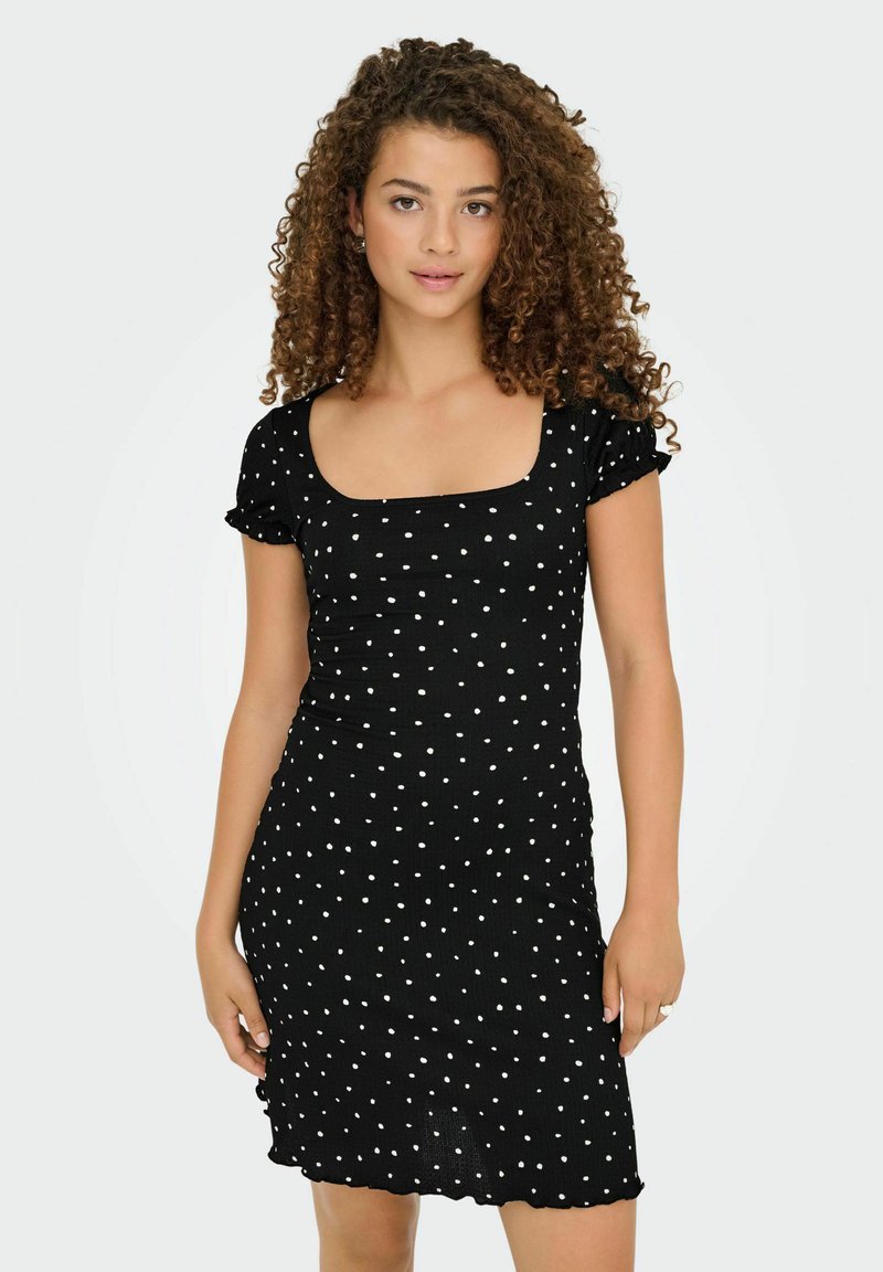 Jeune femme aux cheveux bouclés portant une robe noire à manches courtes et décolleté carré avec des pois blancs, se tenant contre un fond uni.
