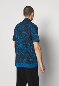 Korte mouwen shirt in blauw met een zwart bladmotief; voorzien van een kraag en een opvallende logoband aan de onderkant. Gladde stofstructuur.