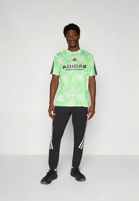 T-shirt desportiva da Adidas com padrão floral verde e logótipo preto, combinada com calças pretas com riscas brancas nas laterais.