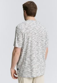 Homme portant un t-shirt à motifs gris clair à manches courtes et un pantalon beige, vu de dos sur un fond uni.