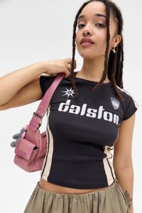 Jauna sieviete ar piedotām matiem, ģērbta melnā "Dalston" logo t-kreklā, khaki svārkos un tur pinku plecu somu ar adītu atslēgu piekariņu.