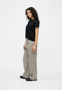 Γυναίκα με σγουρά μαλλιά που φοράει μαύρο μπλουζάκι, μπεζ καρό παντελόνι με φαρδιά πόδια και μαύρα παπούτσια, στέκεται μπροστά σε λευκό φόντο.