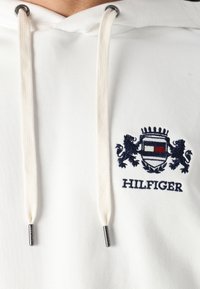 Weiße Kapuzenjacke mit einem marineblauen, gestickten Logo mit Löwen und einem Schild. Die Kordeln sind cremefarben mit Metallakzenten. Der Text lautet "HILFIGER."