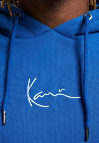 Sudadera con capucha azul hecha de tela suave con un logo blanco bordado. Incluye un cordón con terminales metálicos y una superficie texturizada.