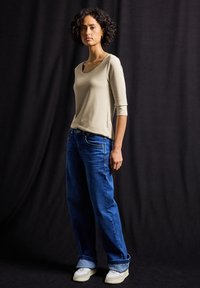 Beżowa bluzka z rękawem 3/4 połączona z luźnymi niebieskimi dżinsami i jasnoskalnymi trampkami, na ciemnym tle.