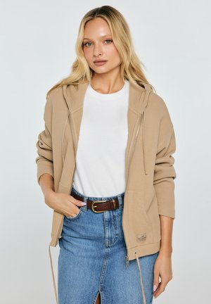 Kobieta ubrana w beżową bluzę na zamek z kapturem na białej koszulce i wysokim stanem niebieską spódnicę dżinsową z brązowym paskiem, stojąca z jedną ręką w kieszeni.