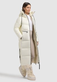Cappotto lungo, imbottito, di un bianco avorio chiaro con finitura lucida, dotato di cappuccio, tasche con zip e dettagli in zip neri a contrasto.