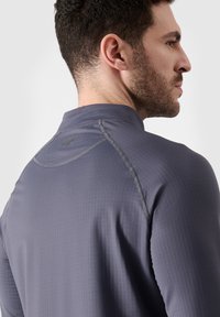 Grijze sportshirt met lange mouwen en hoge kraag, voorzien van textuurstof, contrasterende stiksels en een klein logo op de achterkant.